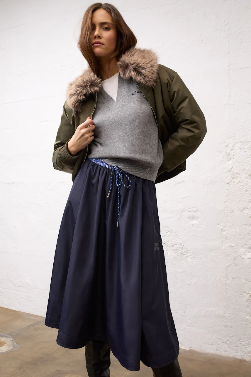 Woman in a navy midi skirt, gray pullover, and green jacket with fur collar, standing confidently.