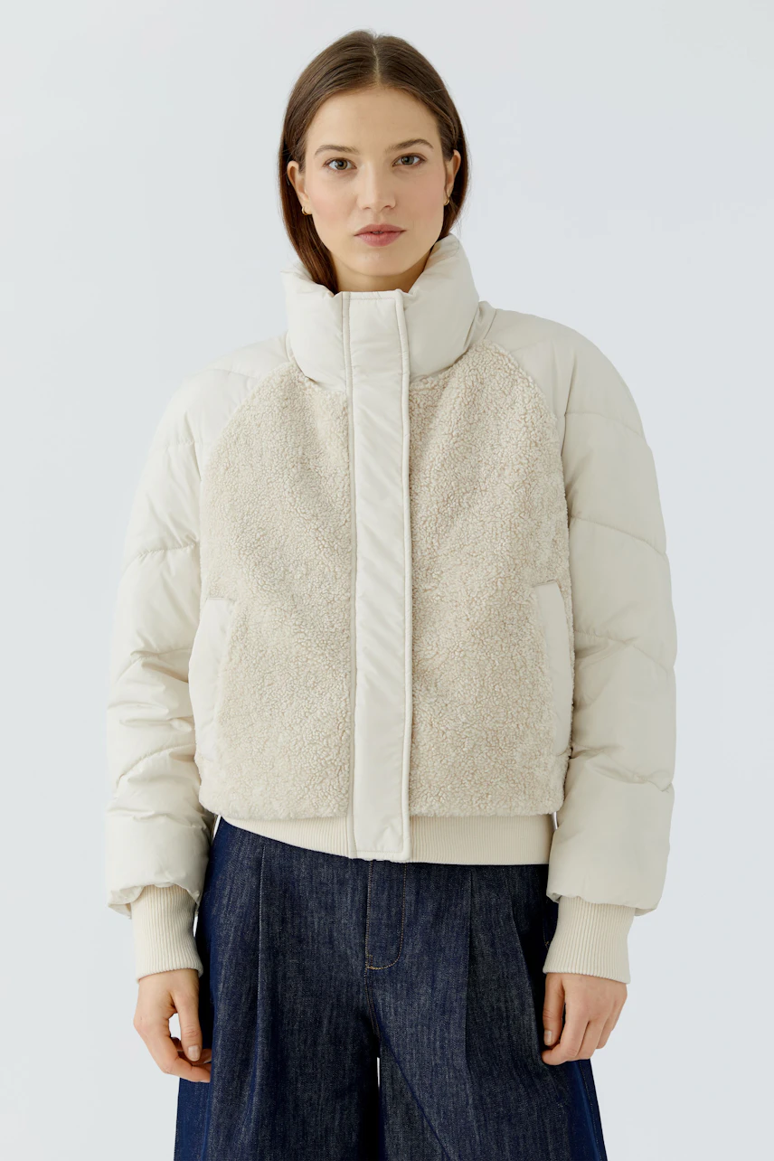 Outdoorjacke: Frau in cremefarbener Jacke mit Stehkragen und flauschigen Details, kombiniert mit Jeans.