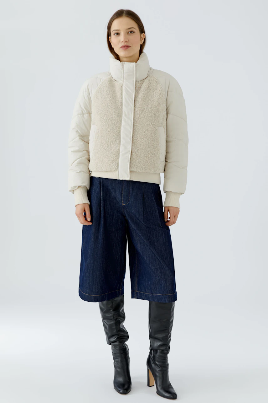 Outdoorjacke: Frau in cremefarbener Jacke mit Stehkragen, kombiniert mit Jeans-Culotte und schwarzen Stiefeln.