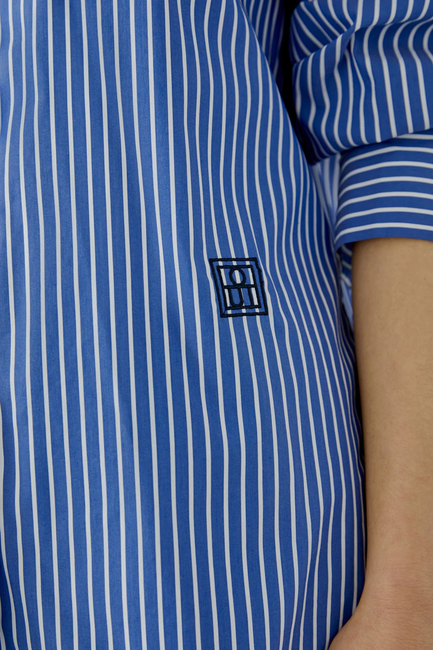 Striped shirt blouse: Close-up of blue and white stripes with logo detail, showcasing casual style.