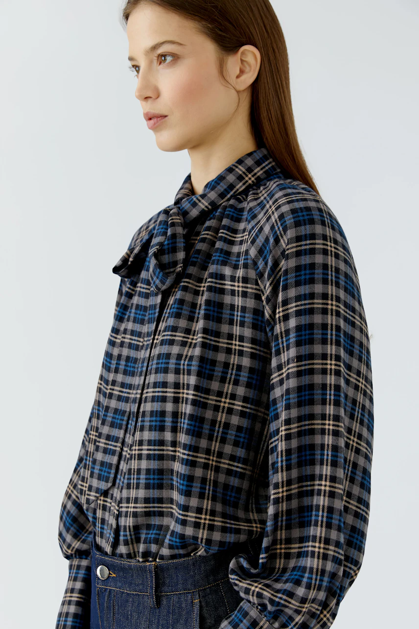 Plaid blouse: Woman in a blue and brown checkered blouse with a bow tie collar, paired with dark jeans.