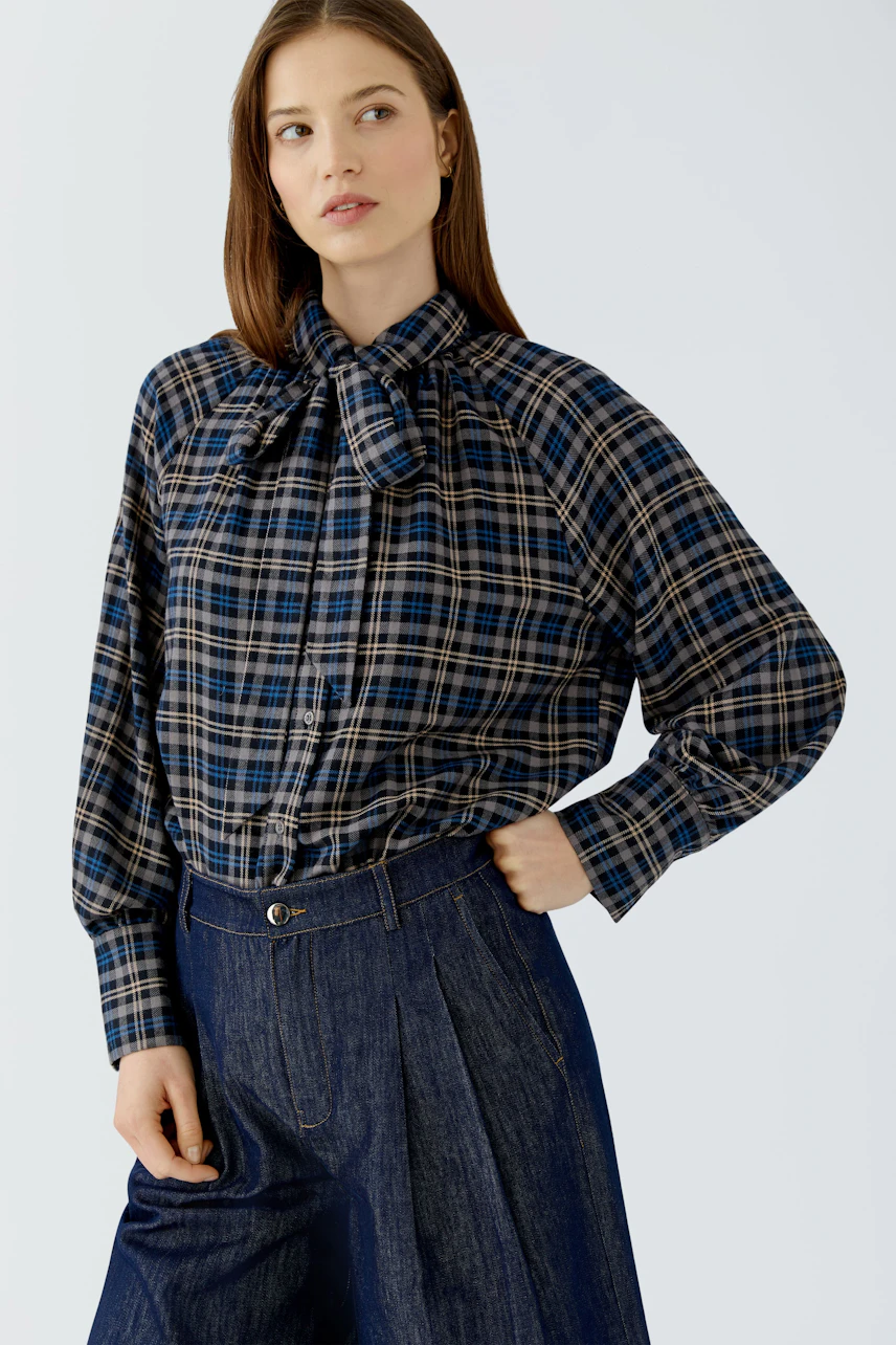 Casual women's blouse: Woman in a plaid blouse with a bow tie collar and relaxed fit, paired with denim pants.