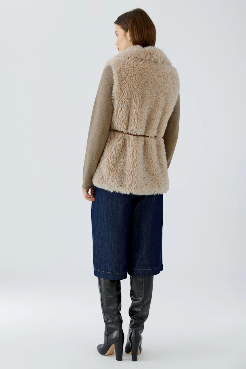 Casual women's vest: Woman in a fluffy beige vest over a brown top, paired with denim culottes and tall boots.
