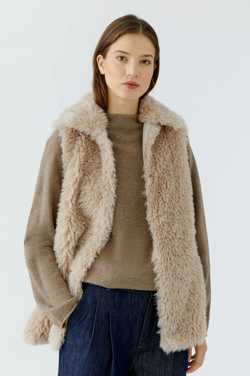 Casual women's vest: Young woman in a fluffy beige vest over a brown pullover, paired with blue jeans.