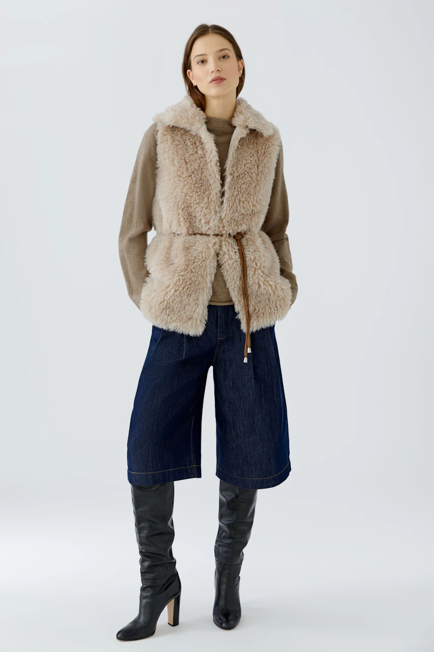 Casual women's vest: Woman in beige fluffy vest over brown top, denim culottes, and knee-high boots.