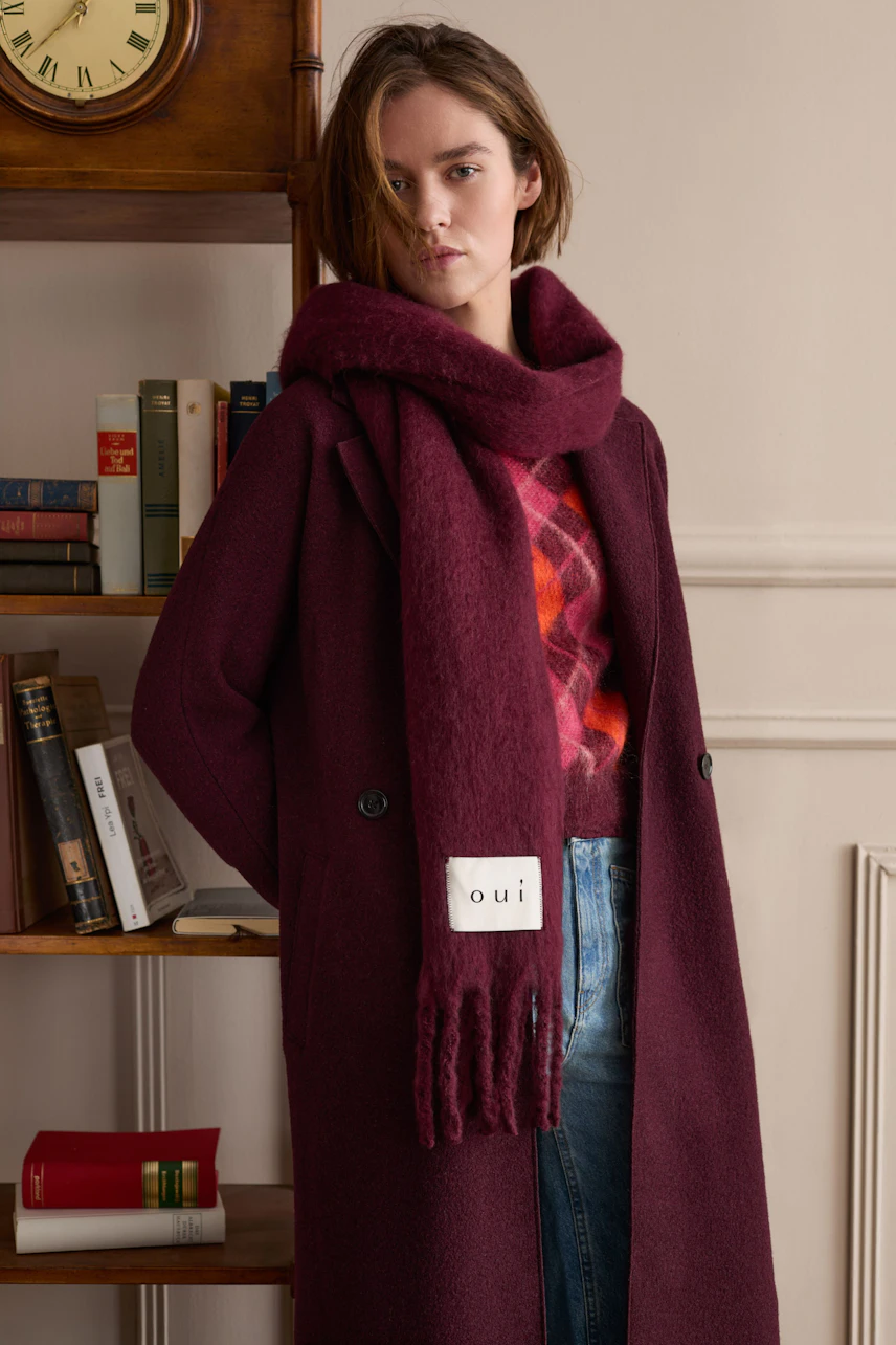 Frau im burgunderroten Mantel mit passendem Schal, darunter ein karierter Pullover. Lässiger Look.
