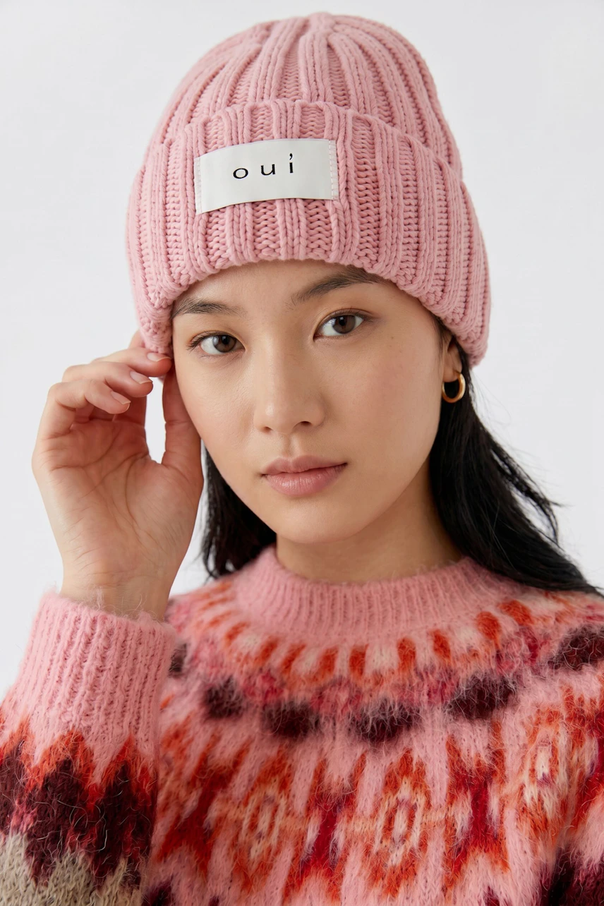 Woman in pink knit hat with 'oui' logo, wearing a colorful patterned sweater, looking at the camera.