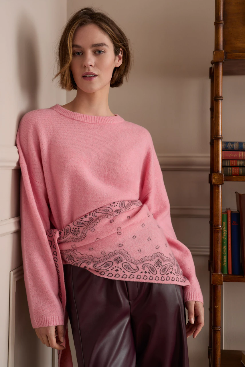 Frau im rosa Pullover mit Paisley-Muster, kombiniert mit brauner Hose, steht lässig an der Wand.