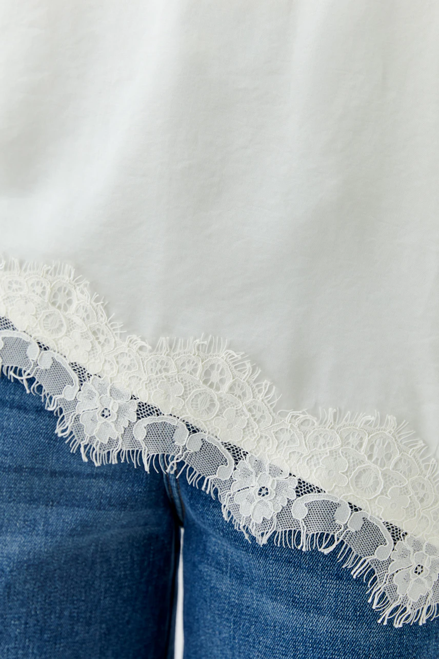 Weißes Damen-Top mit Spitzenbesatz am Saum, kombiniert mit blauer Jeans. Schlicht und stilvoll.