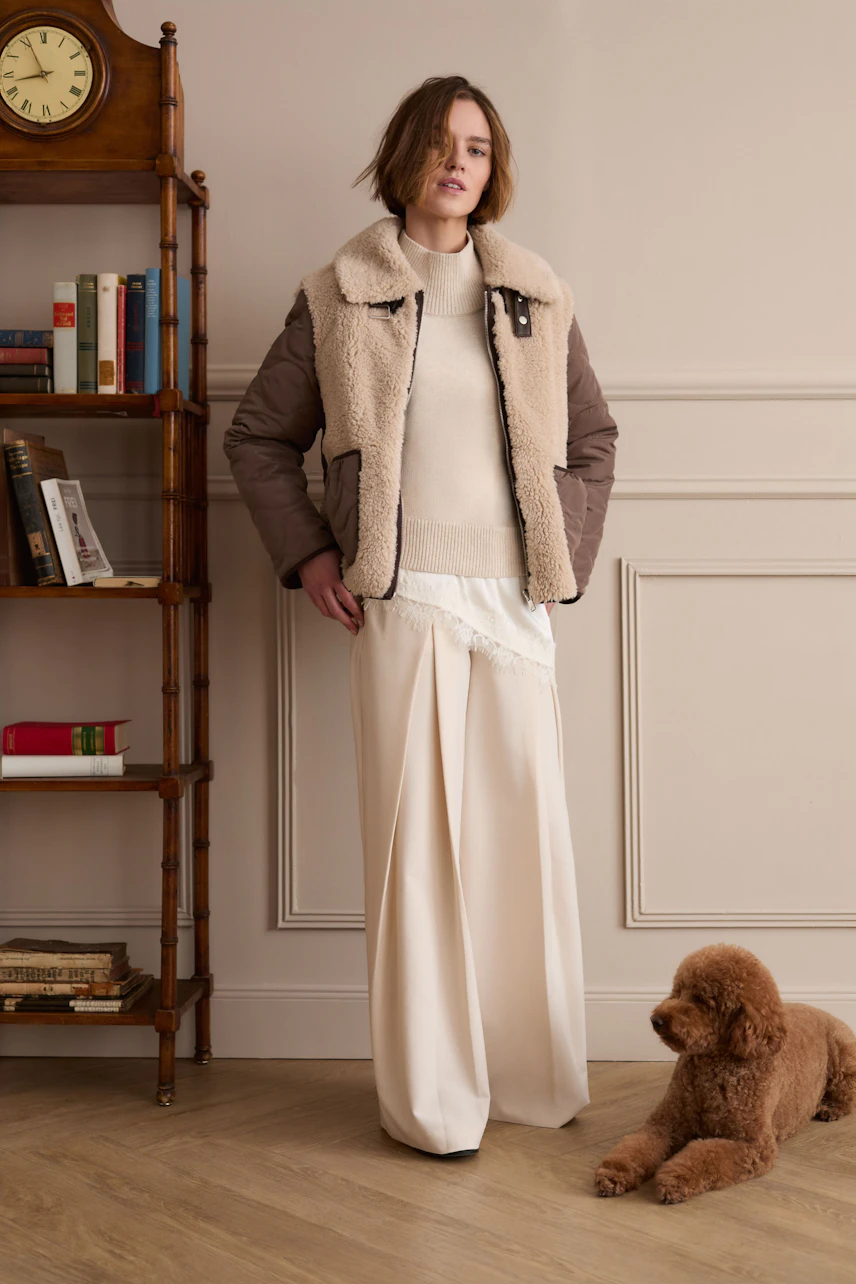Frau im cremefarbenen Top mit beiger Jacke und weiter Hose, steht neben einem Hund in einem stilvollen Raum.