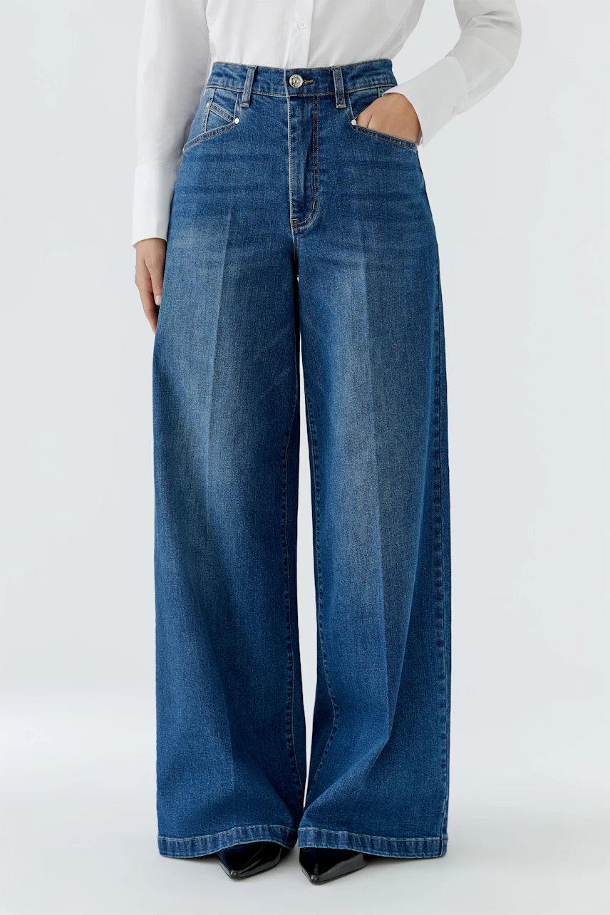 Jeans Wide Leg: Frau in blauer, weit geschnittener Jeans mit weißer Bluse und schwarzen Schuhen.