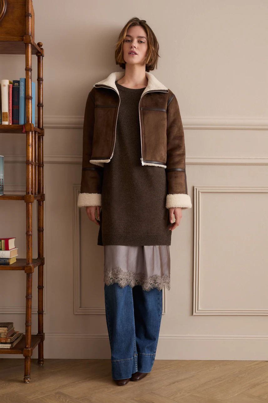 Woman in flared jeans, layered dress, and brown shearling jacket stands confidently.