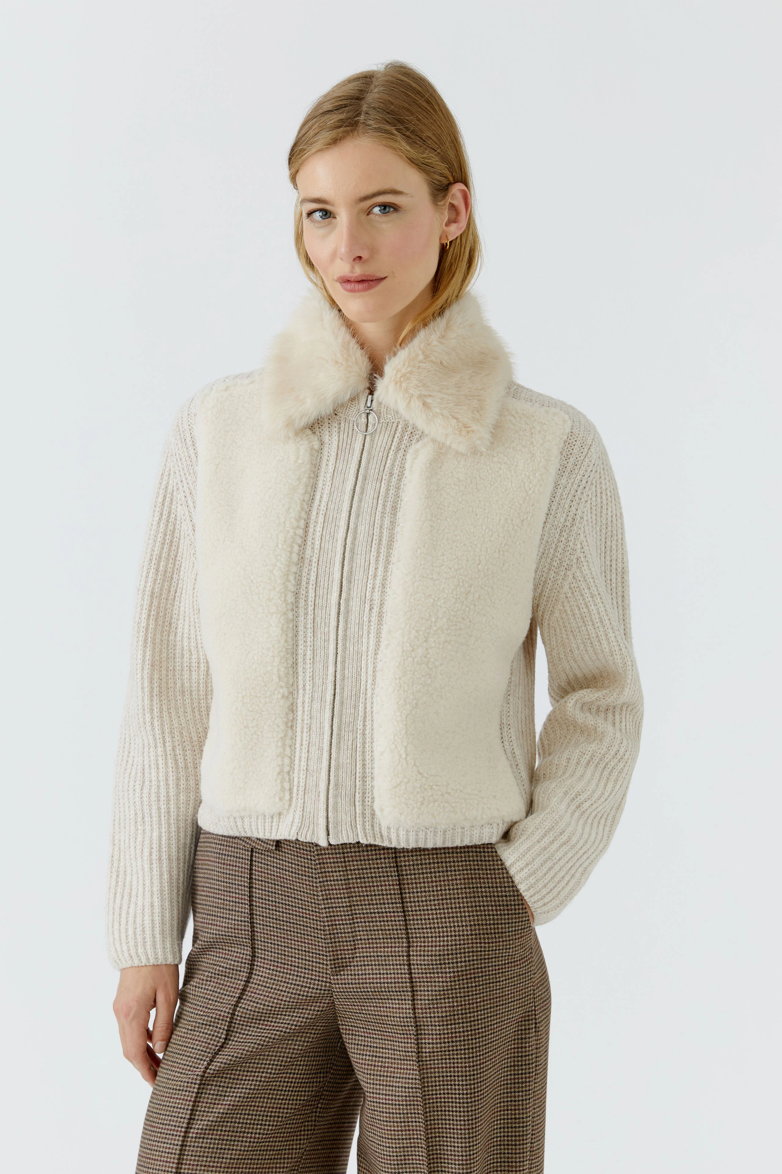 Strickjacke: Frau in cremefarbener Strickjacke mit Fellkragen, kombiniert mit karierter Hose.