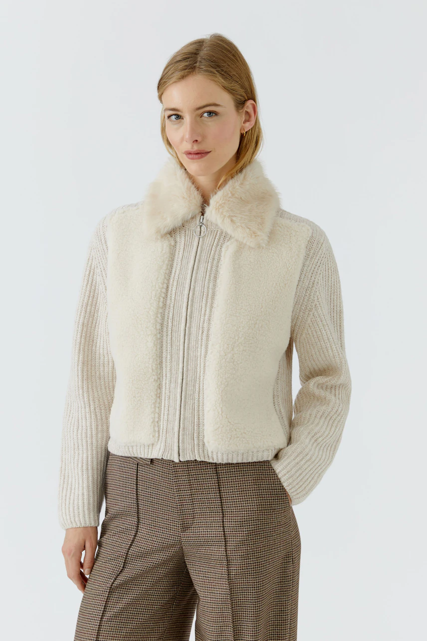 Strickjacke: Frau in cremefarbener Strickjacke mit Fellkragen, kombiniert mit karierter Hose.