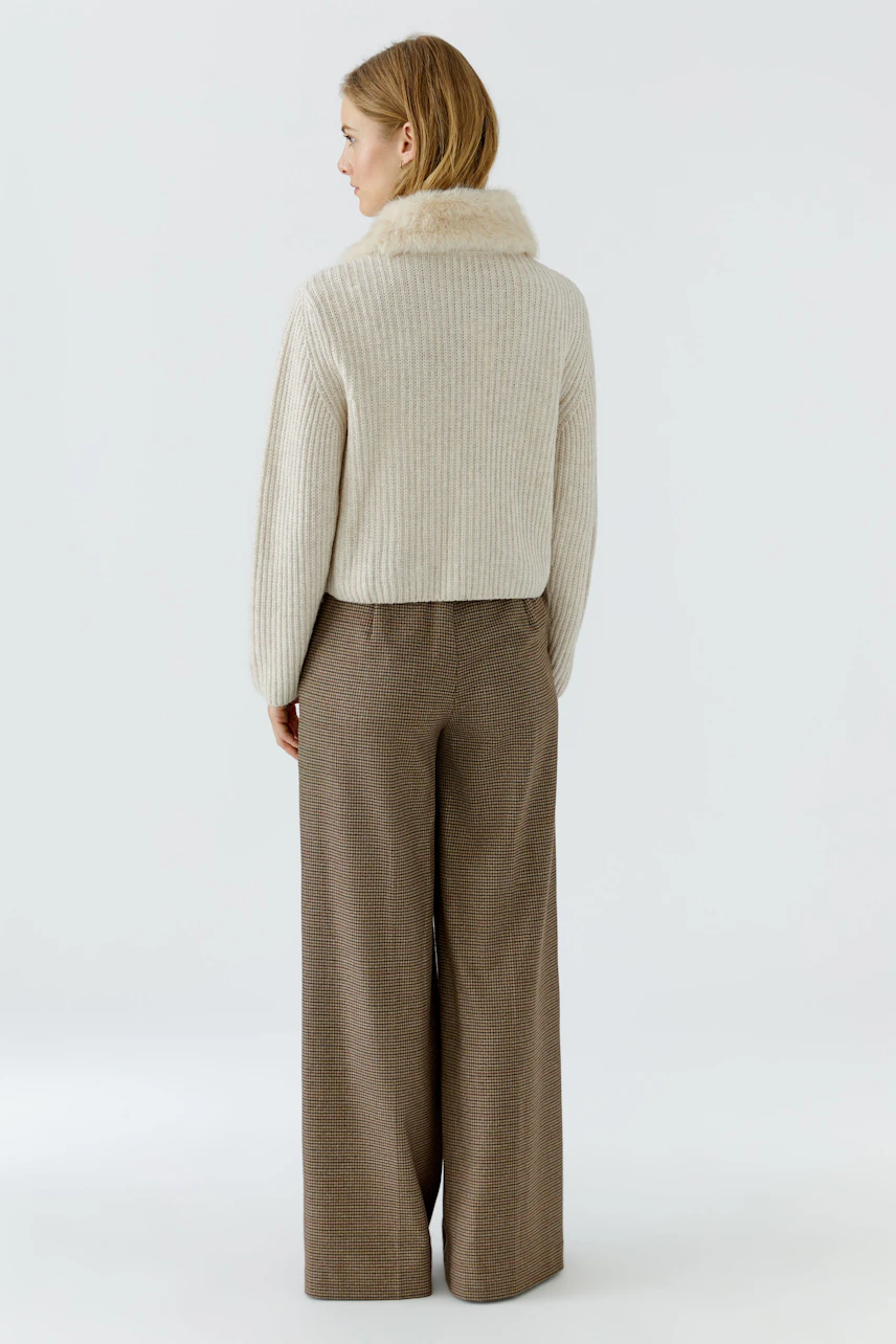 Frau in beiger Strickjacke mit hohem Kragen, kombiniert mit brauner Hose. Rückansicht.
