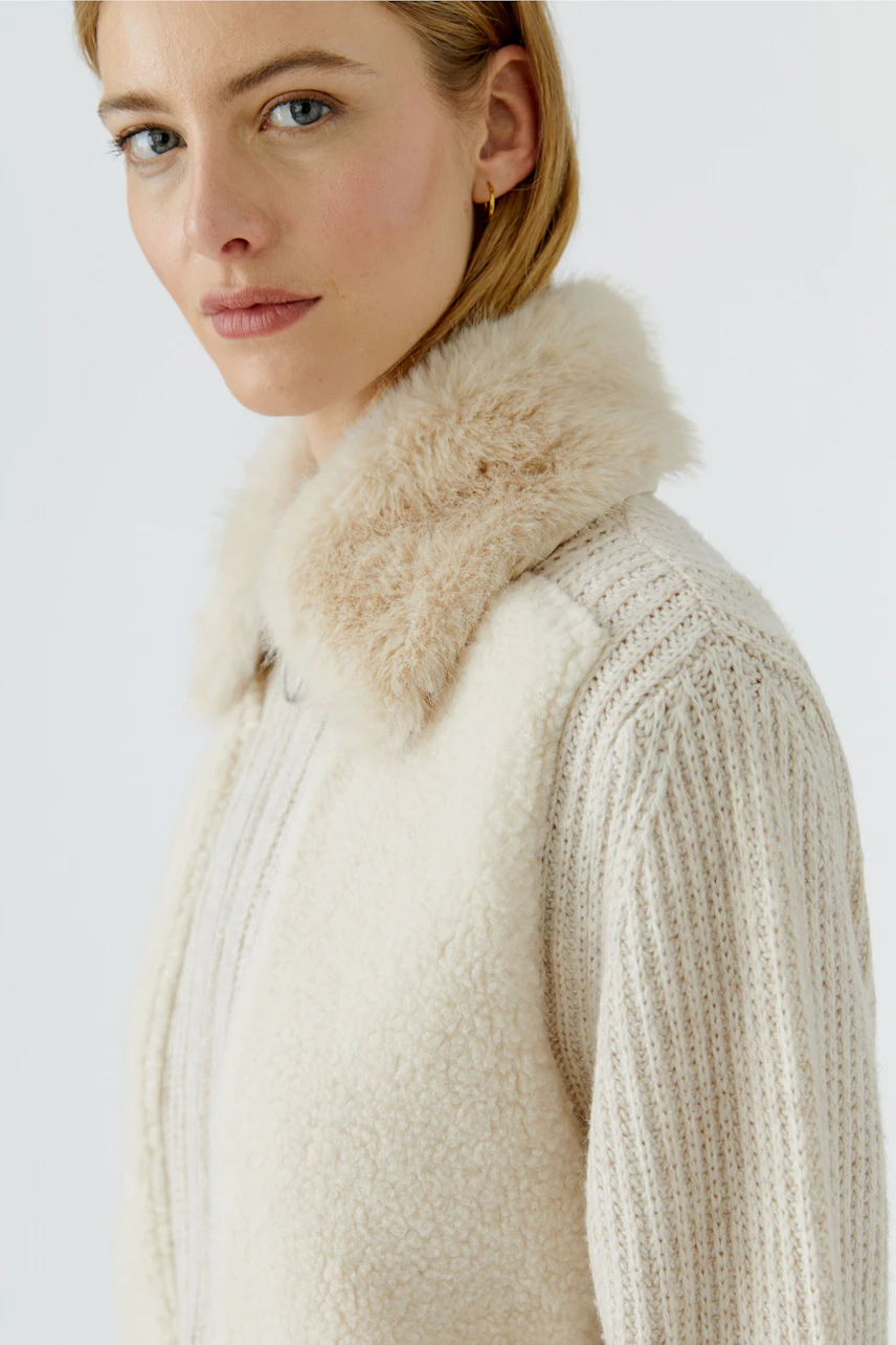 Frau in cremefarbener Strickjacke mit flauschigem Kragen, schaut zur Seite.