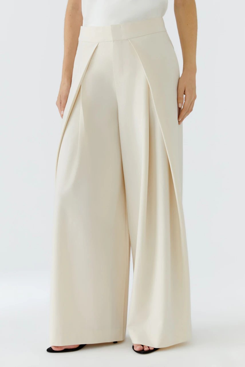 Woman in cream pleated trousers, standing confidently. Wide-leg design with a high waist.