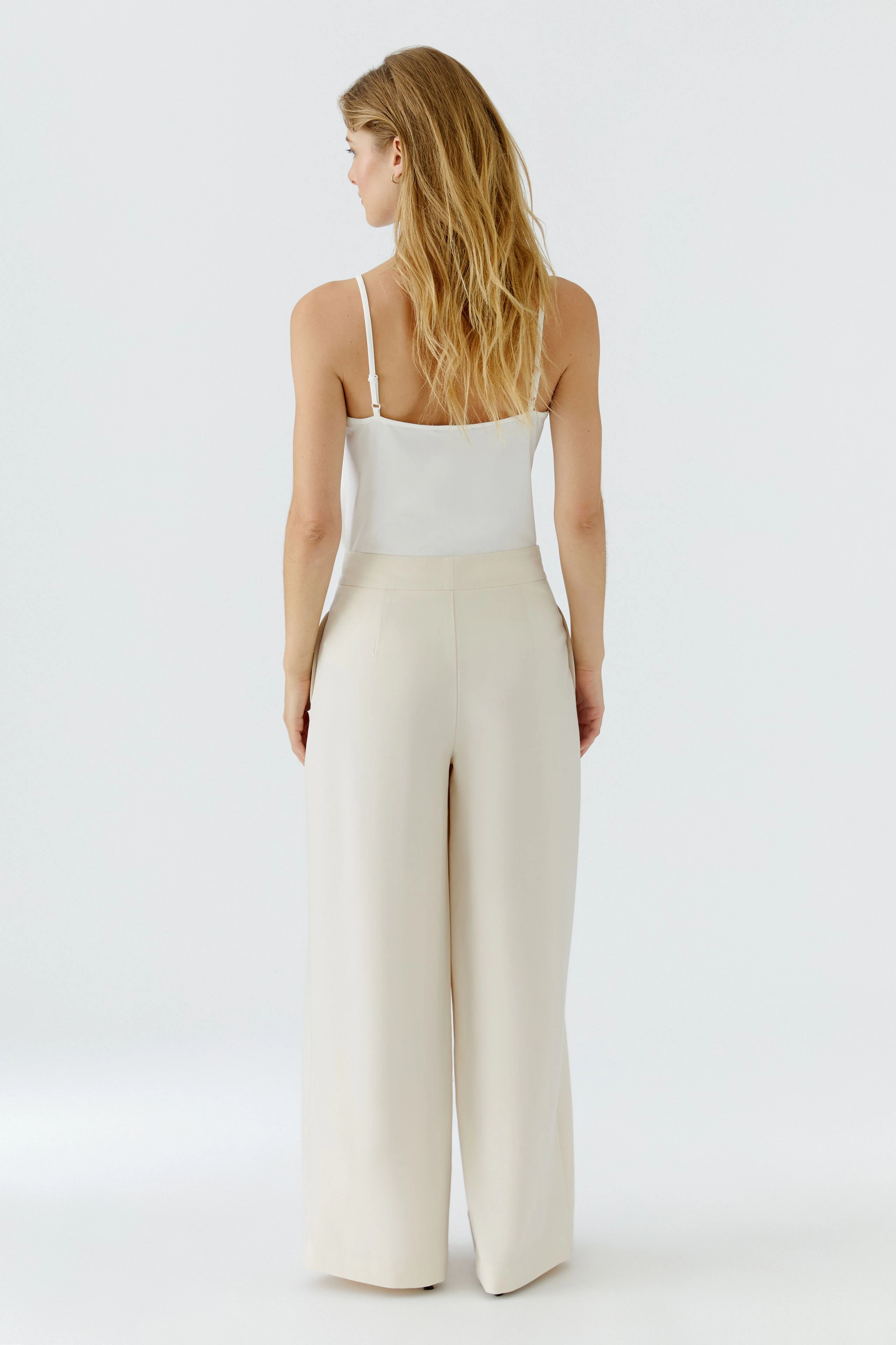 Frau in weißer Bundfaltenhose und Top, Rückansicht. Die Hose ist weit geschnitten und modern.