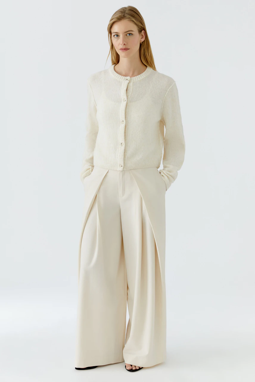 Woman in wide pleated trousers and white cardigan, hands in pockets, standing confidently.