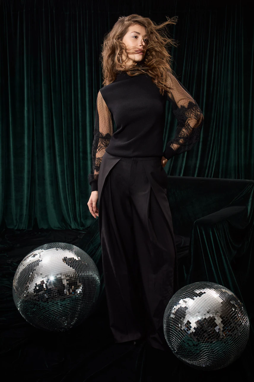 Woman in black pleated trousers and lace-sleeve top, standing confidently with disco balls.