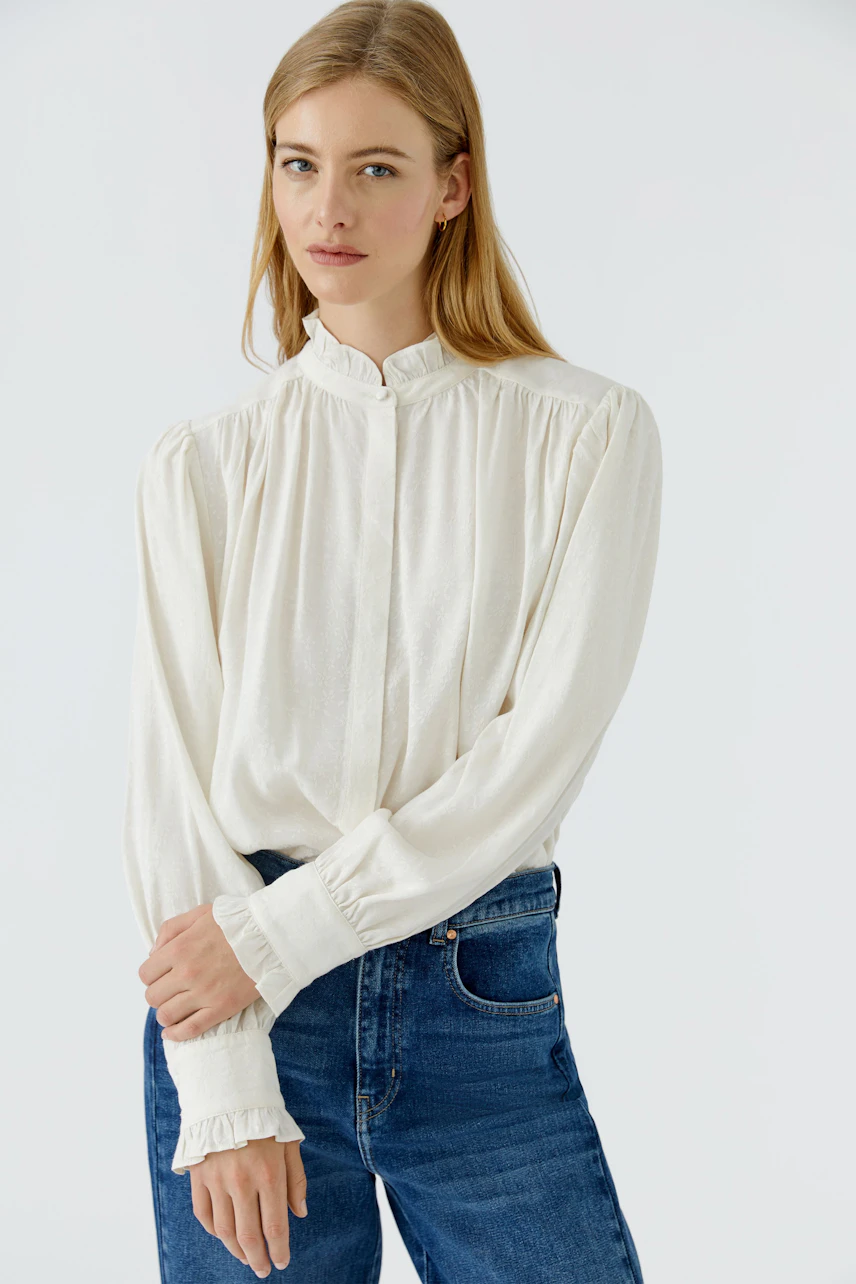Casual women's blouse: Woman in a white blouse with ruffled cuffs and blue jeans, looking relaxed.