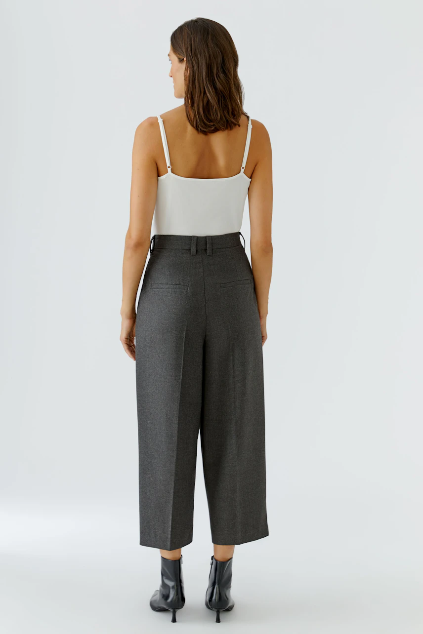 Frau in grauer Bundfaltenhose, weißem Top und Stiefeletten. Rückansicht zeigt lässigen, modernen Look.
