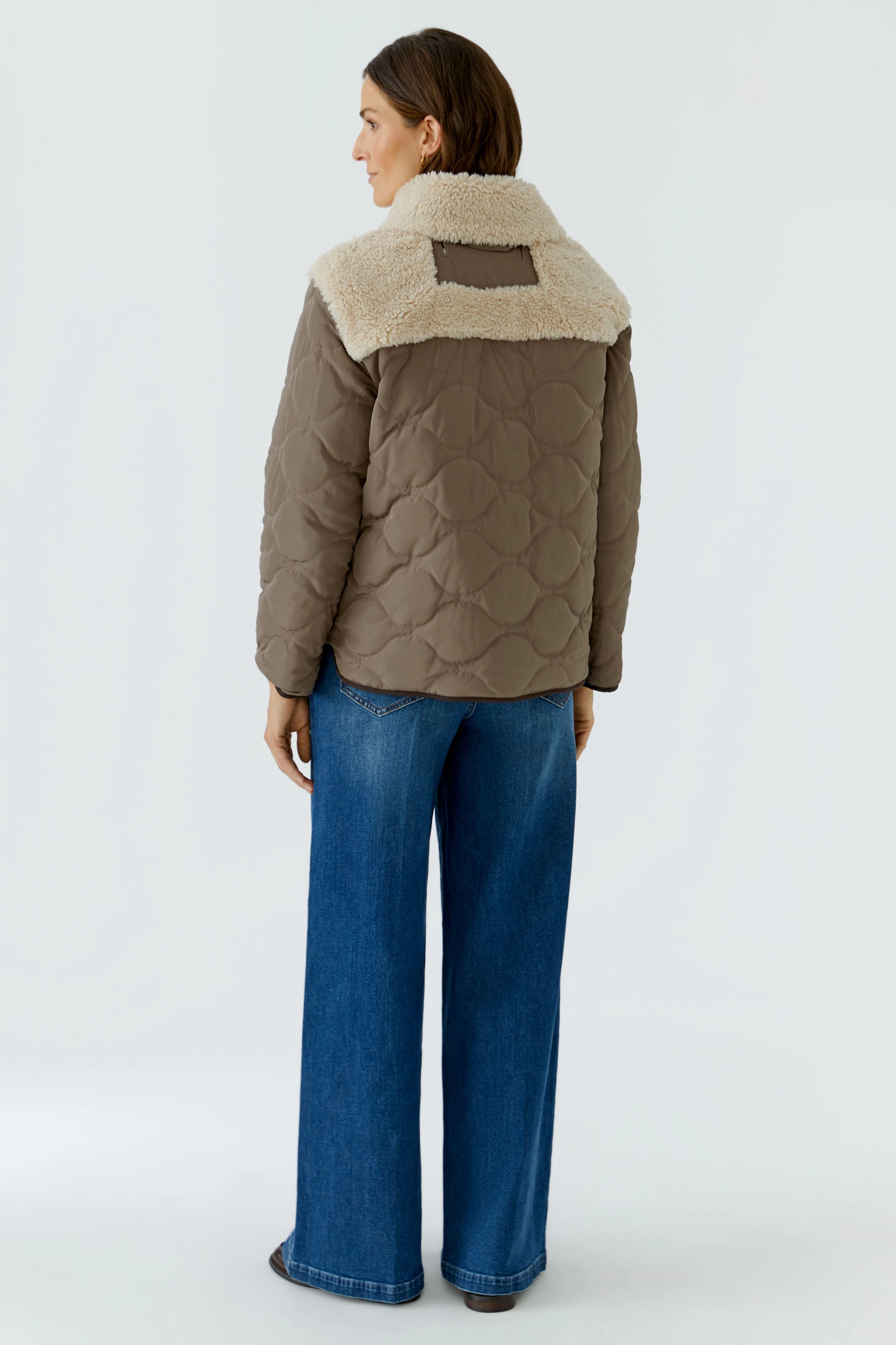 Outdoorjacke: Frau in brauner Steppjacke mit hellem Kragen, kombiniert mit blauer Jeans, von hinten.