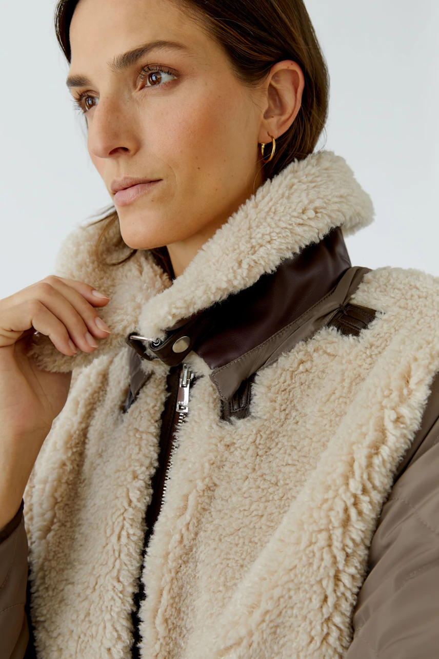 Woman in cozy outdoor jacket with shearling collar, brown leather accents, and zip closure, looking thoughtful.