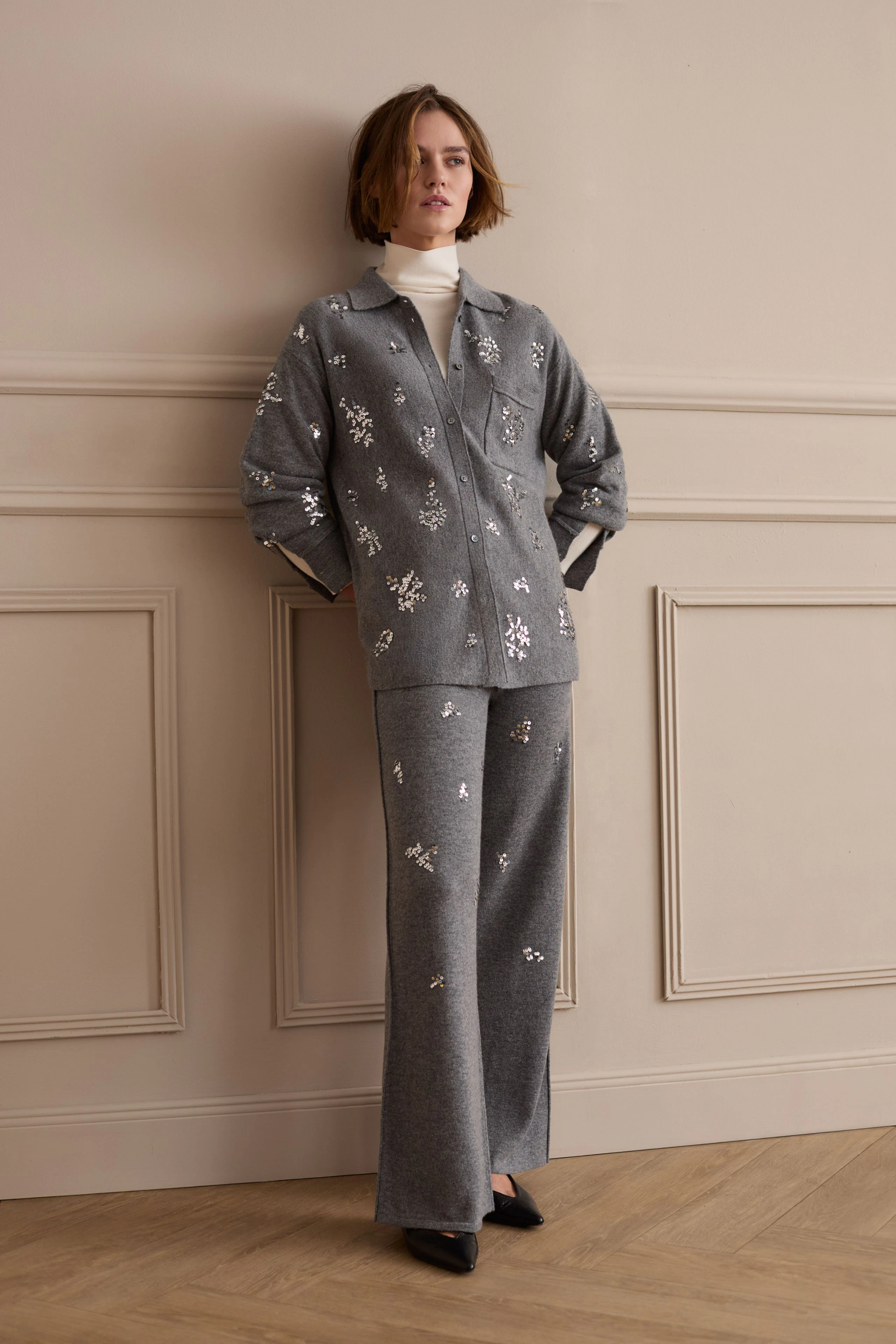 Woman in grey knit pants with floral patterns, matching top, and white turtleneck, leaning against wall.