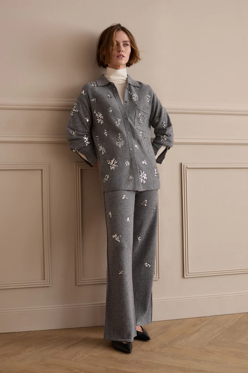 Strickhose: Frau in grauer Strickhose mit floralen Mustern, kombiniert mit passender Jacke, steht lässig an der Wand.