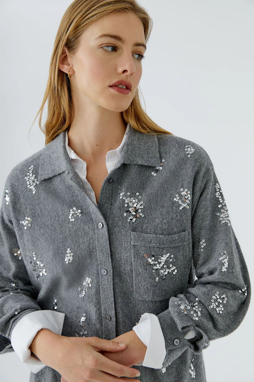 Graue Strickjacke: Frau trägt graue Strickjacke mit funkelnden Details, darunter weiße Bluse.