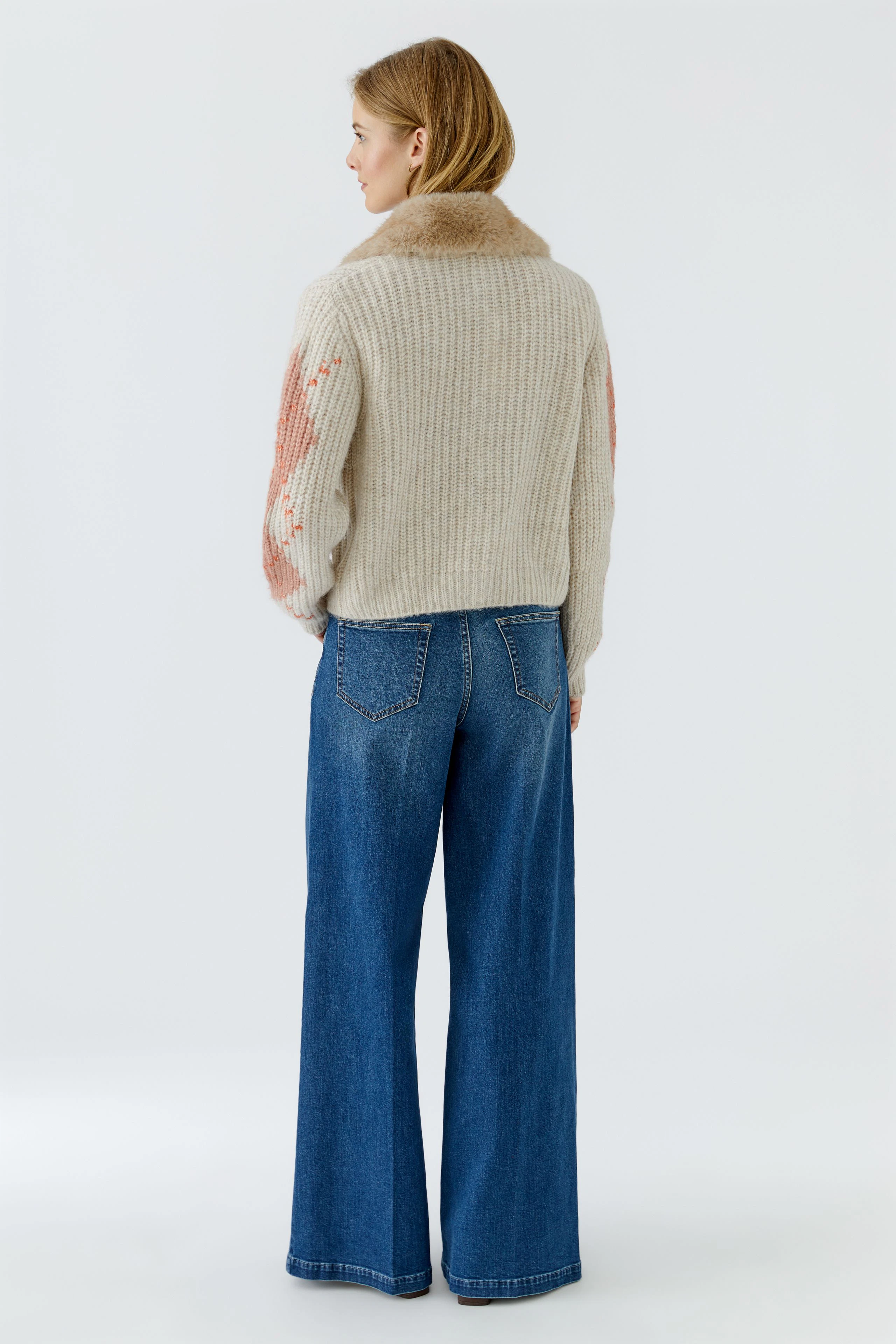 Strickjacke: Frau in beiger Strickjacke mit Muster, kombiniert mit blauer Jeans, von hinten zu sehen.