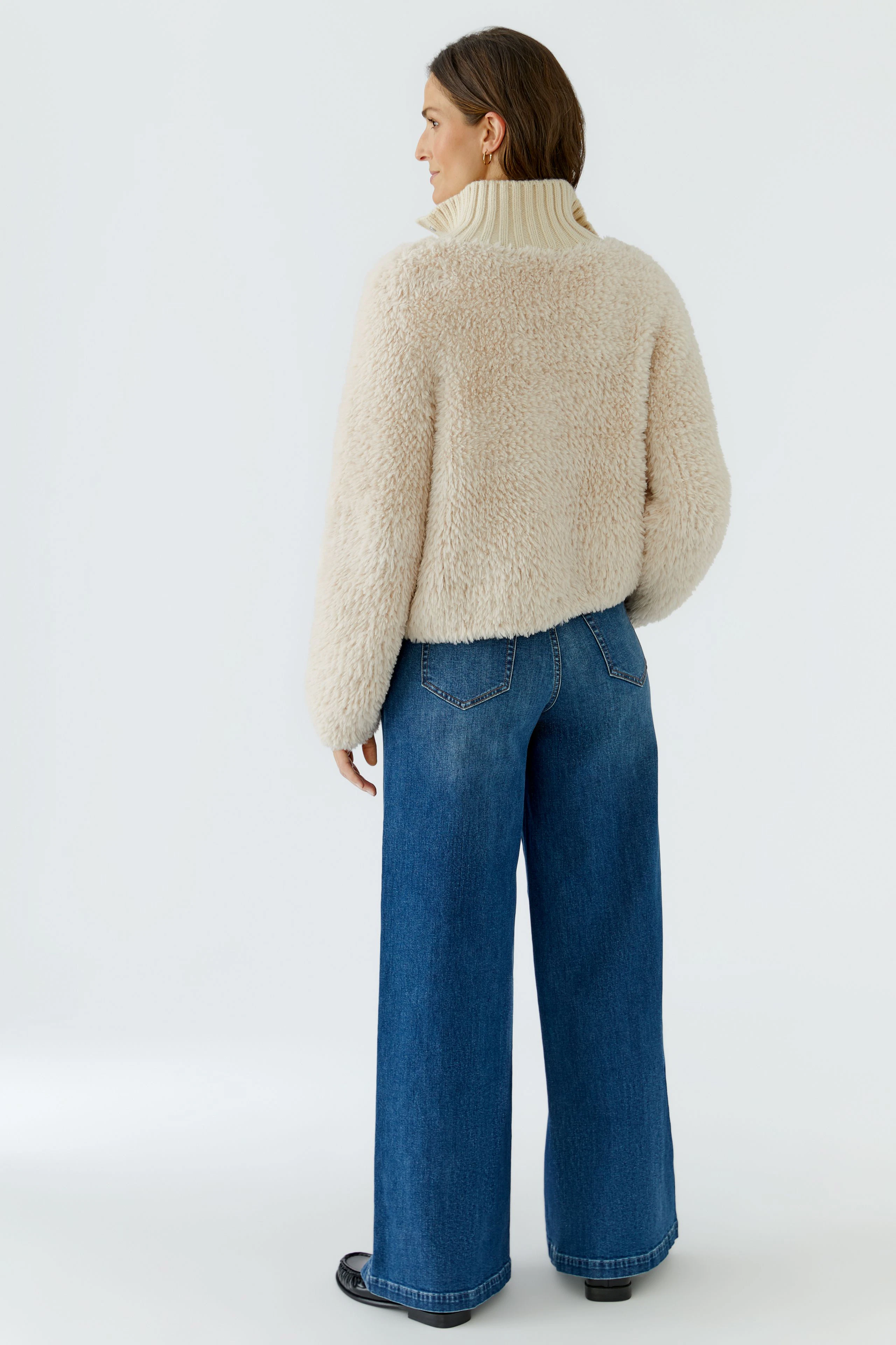 Frau in beiger Strickjacke mit hohem Kragen, kombiniert mit blauer Jeans, von hinten gesehen.