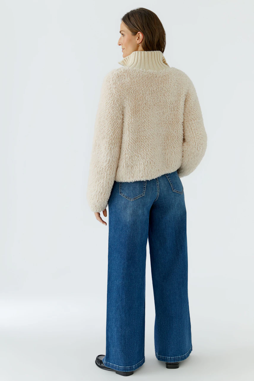 Woman in cozy cardigan: Beige knit cardigan with high collar, paired with wide-leg jeans, viewed from the back.