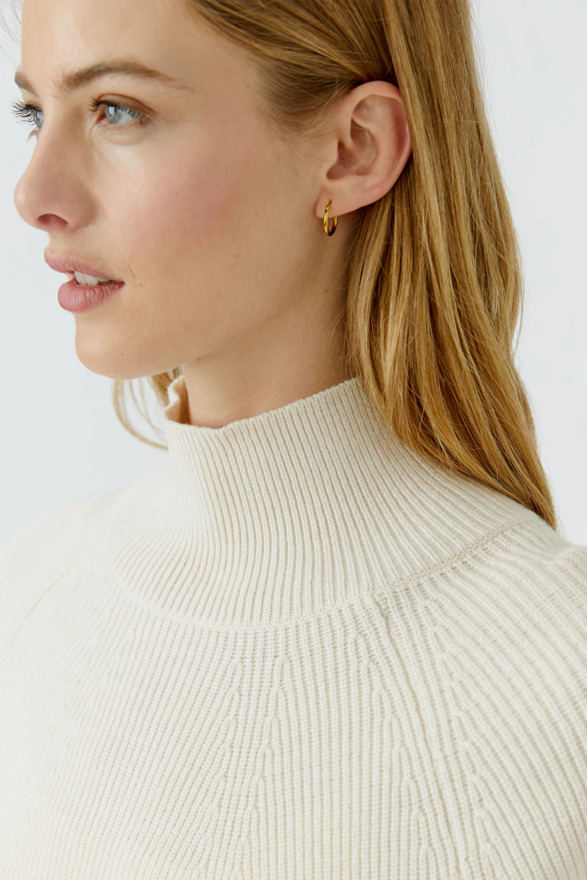 Woman in a cream ribbed pullover with a high neck, looking to the side. Simple and stylish design.