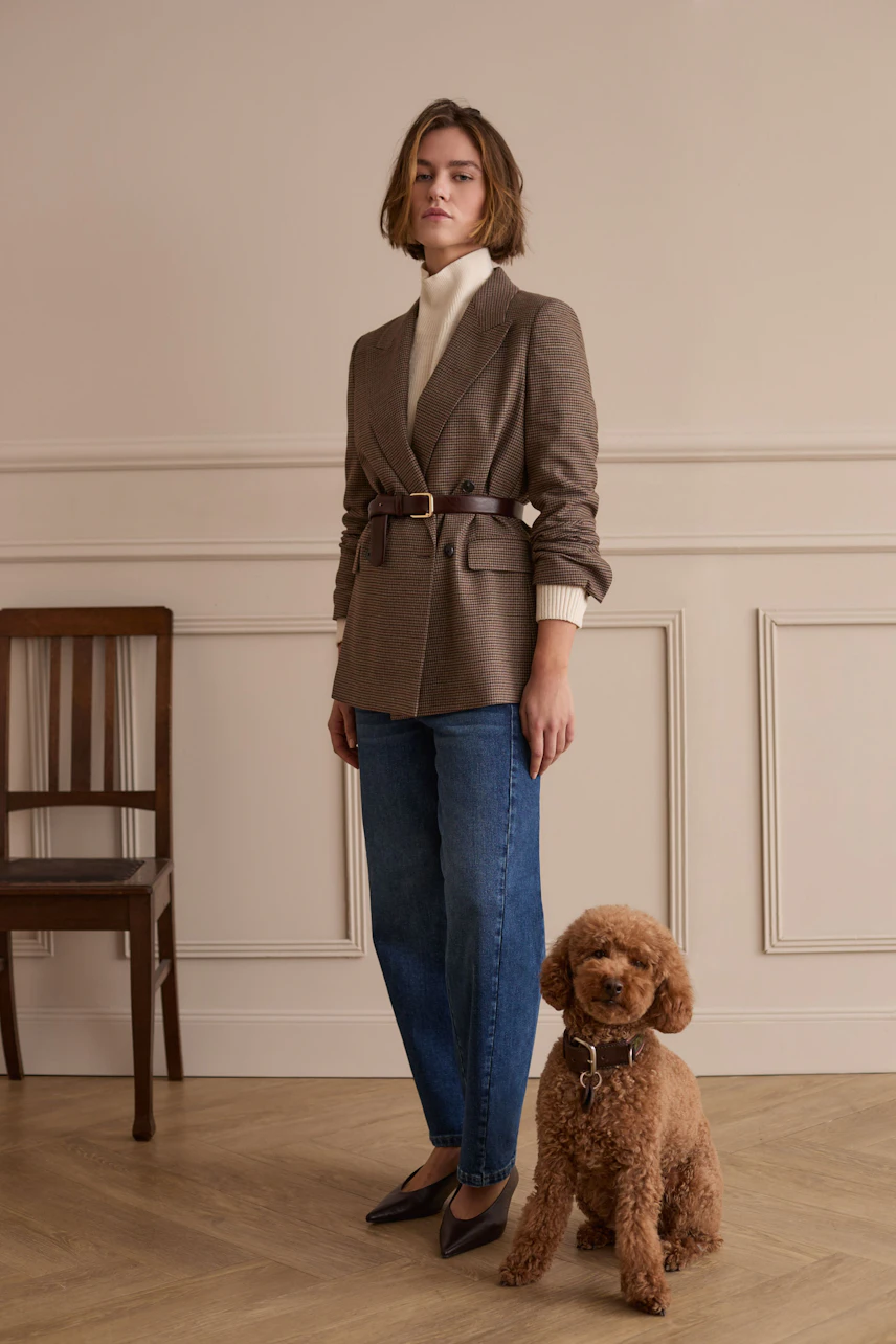 Woman in a beige blazer over a white pullover, paired with blue jeans, standing confidently with a dog.