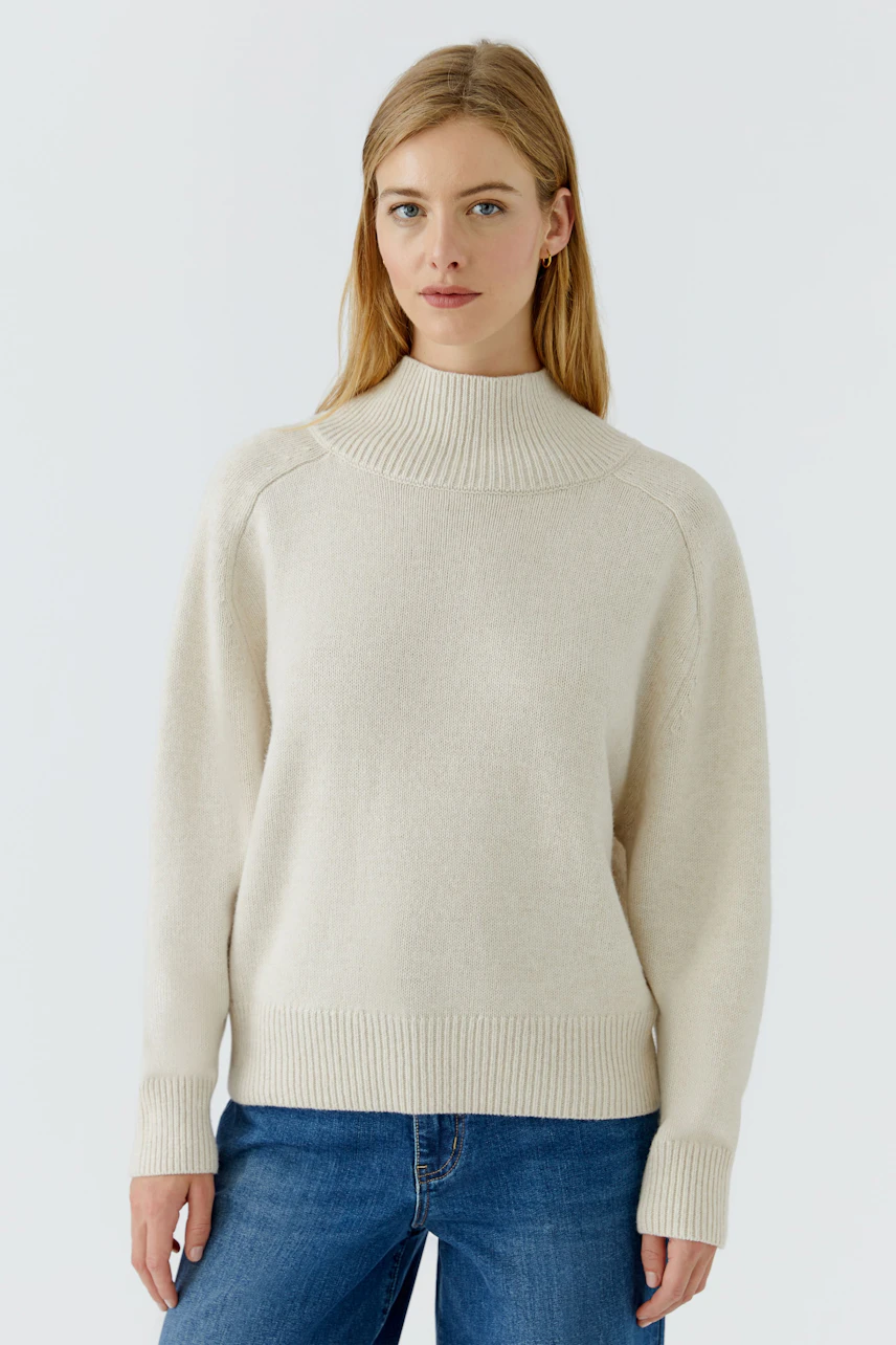Frau im cremefarbenen Pullover mit Stehkragen und blauer Jeans, schlicht und bequem.