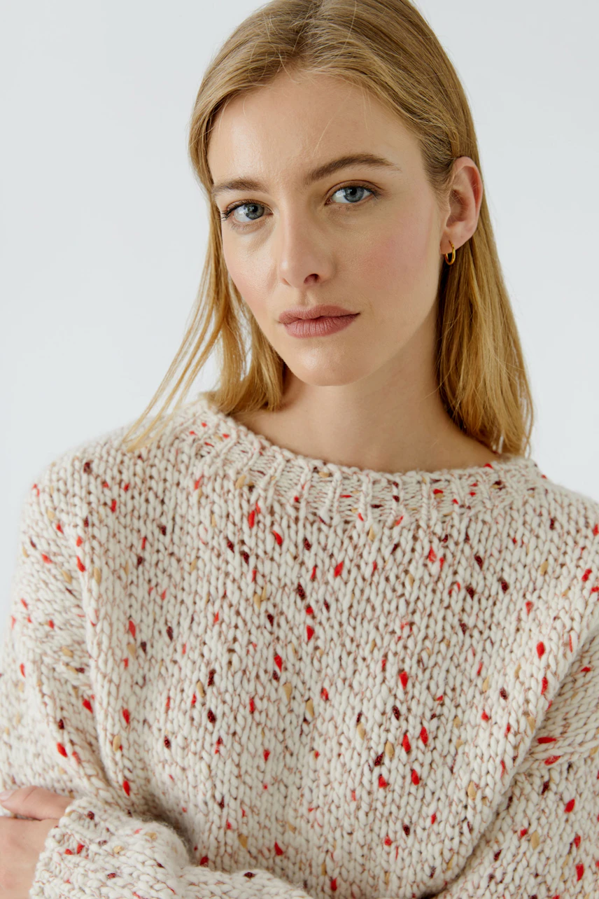 Frau im grob gestrickten Pullover mit roten Akzenten, lässig und modern, schaut direkt in die Kamera.