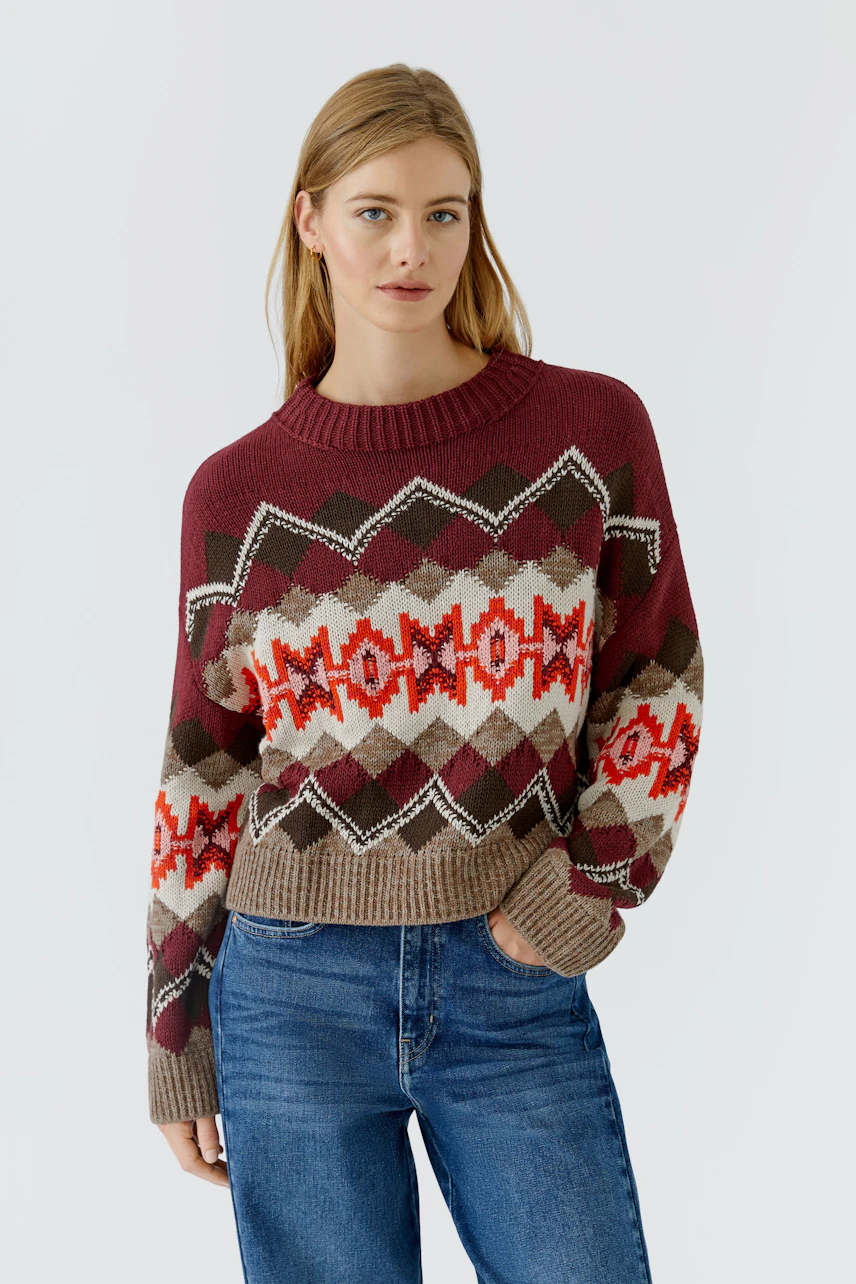Frau im gemusterten Pullover: Roter Strickpullover mit geometrischem Muster, kombiniert mit blauer Jeans.