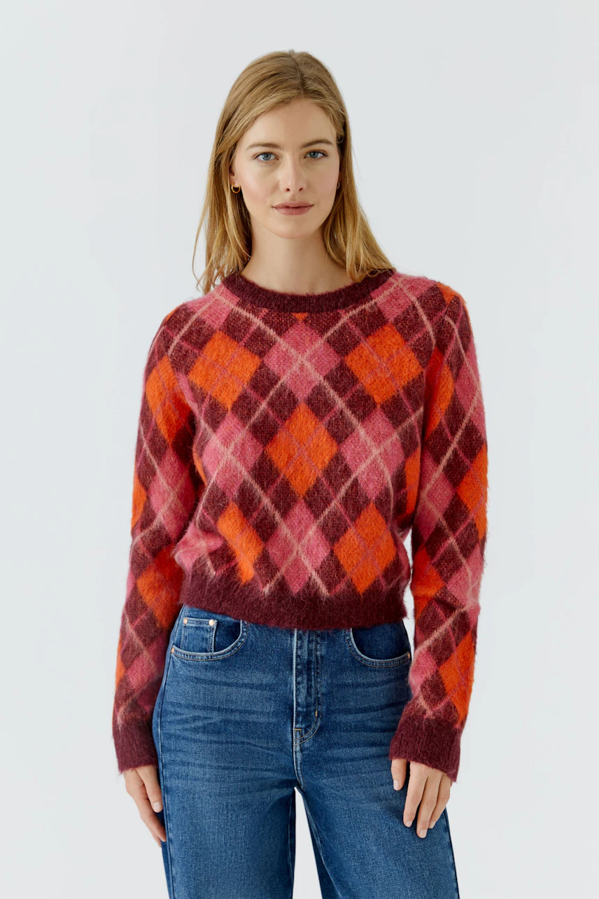 Frau im karierten Pullover in Rot- und Pinktönen, kombiniert mit blauen Jeans, lässig und modern.