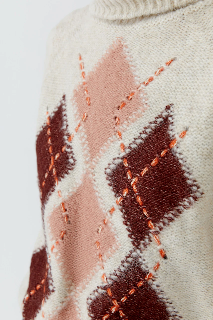 Gemusterter Damen-Pullover: Nahaufnahme eines cremefarbenen Pullovers mit geometrischem Muster in Rot- und Rosatönen.
