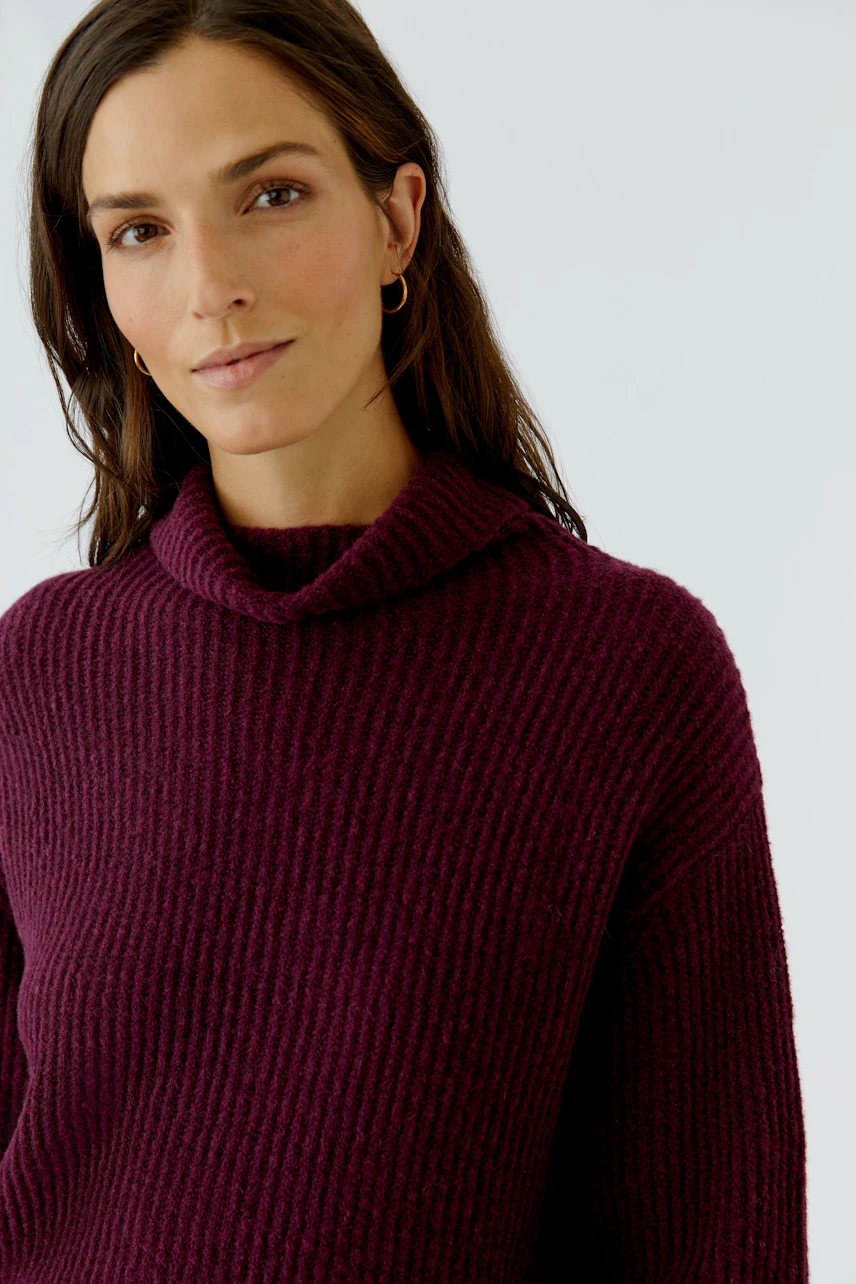 Frau im weinroten Pullover mit Rollkragen, lässig und modern, schaut freundlich in die Kamera.