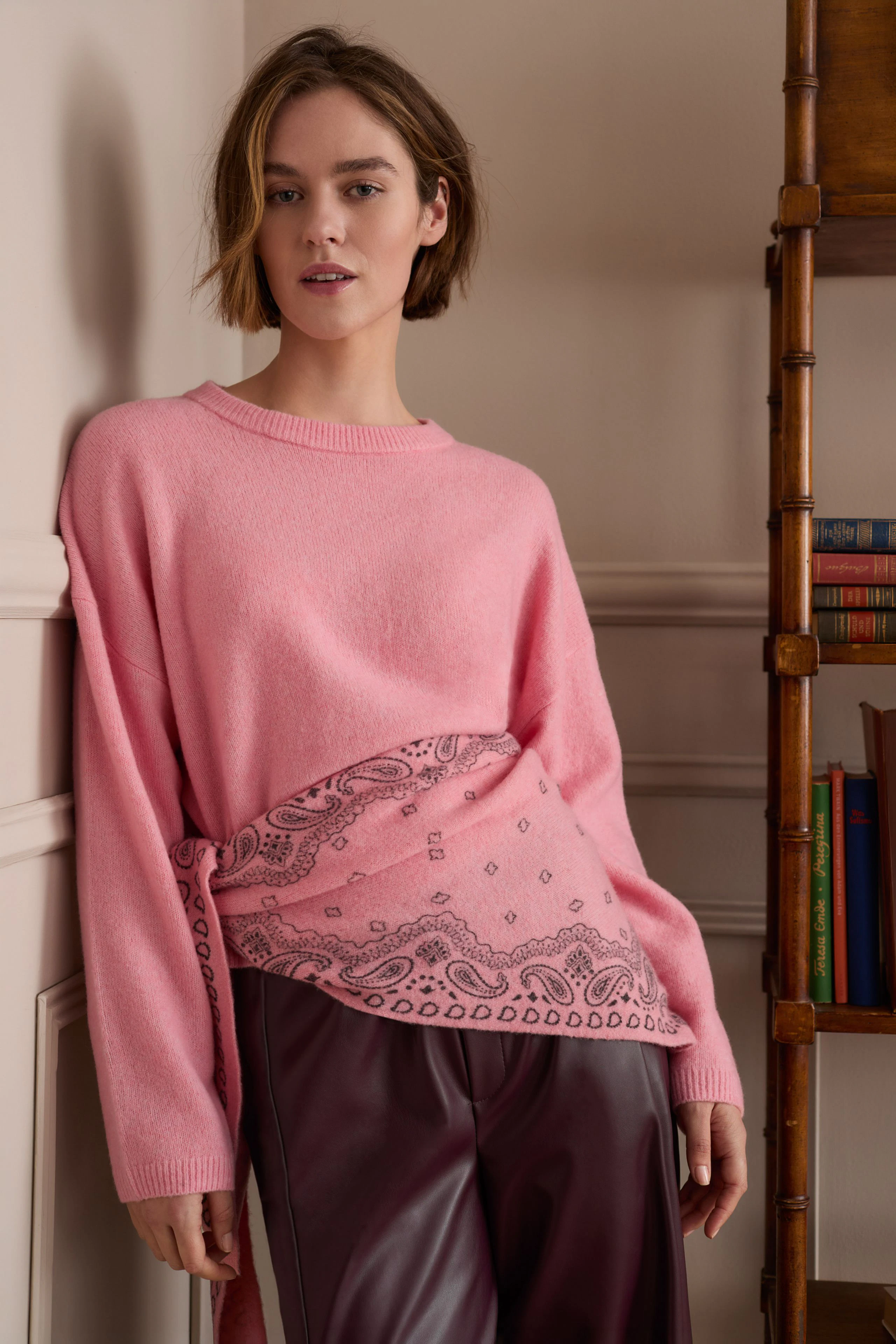 Woman in pink pullover with paisley scarf, paired with maroon pants, standing confidently indoors.