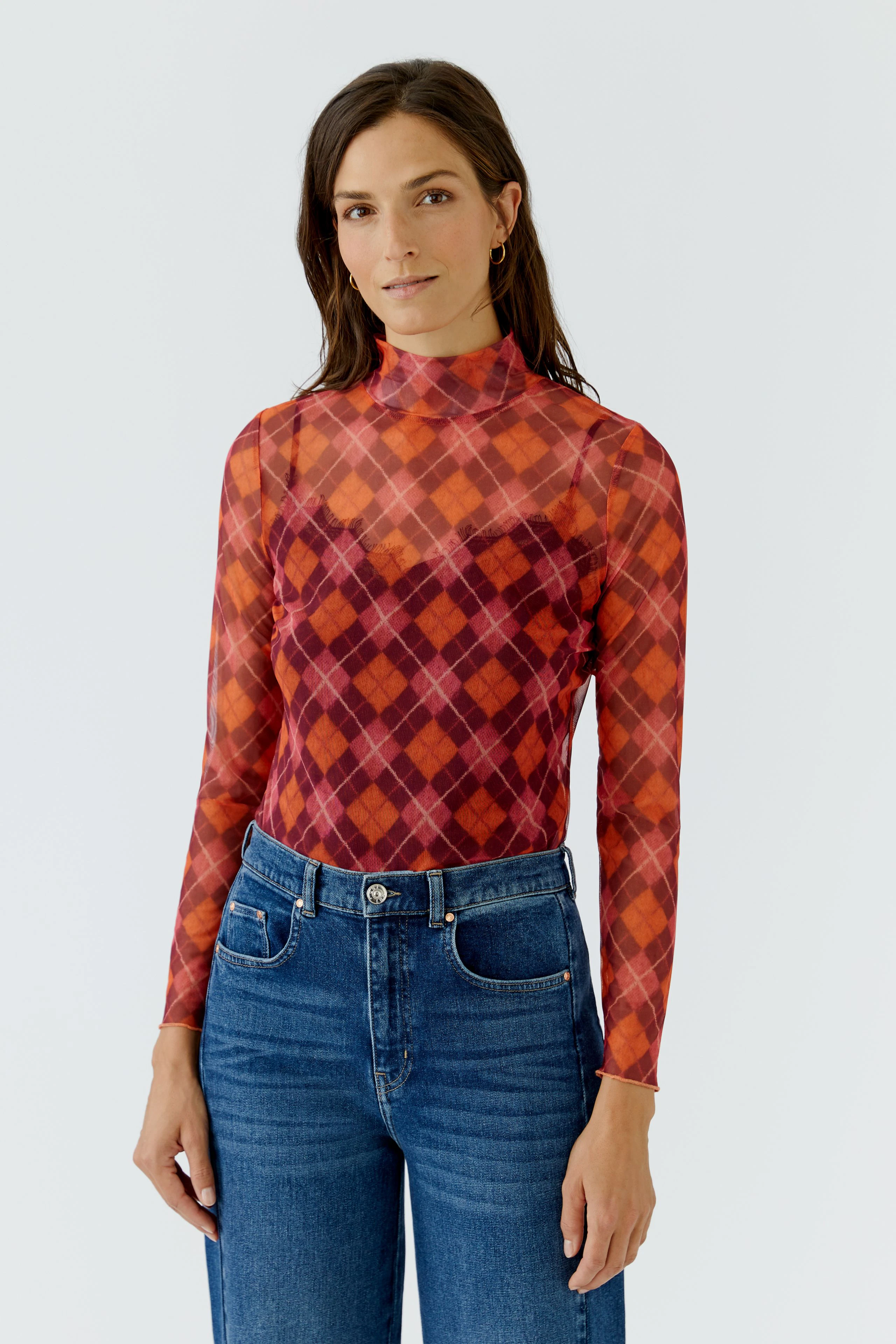 Frau im karierten Langarmshirt in Rot- und Orangetönen, kombiniert mit blauen Jeans, schaut direkt in die Kamera.