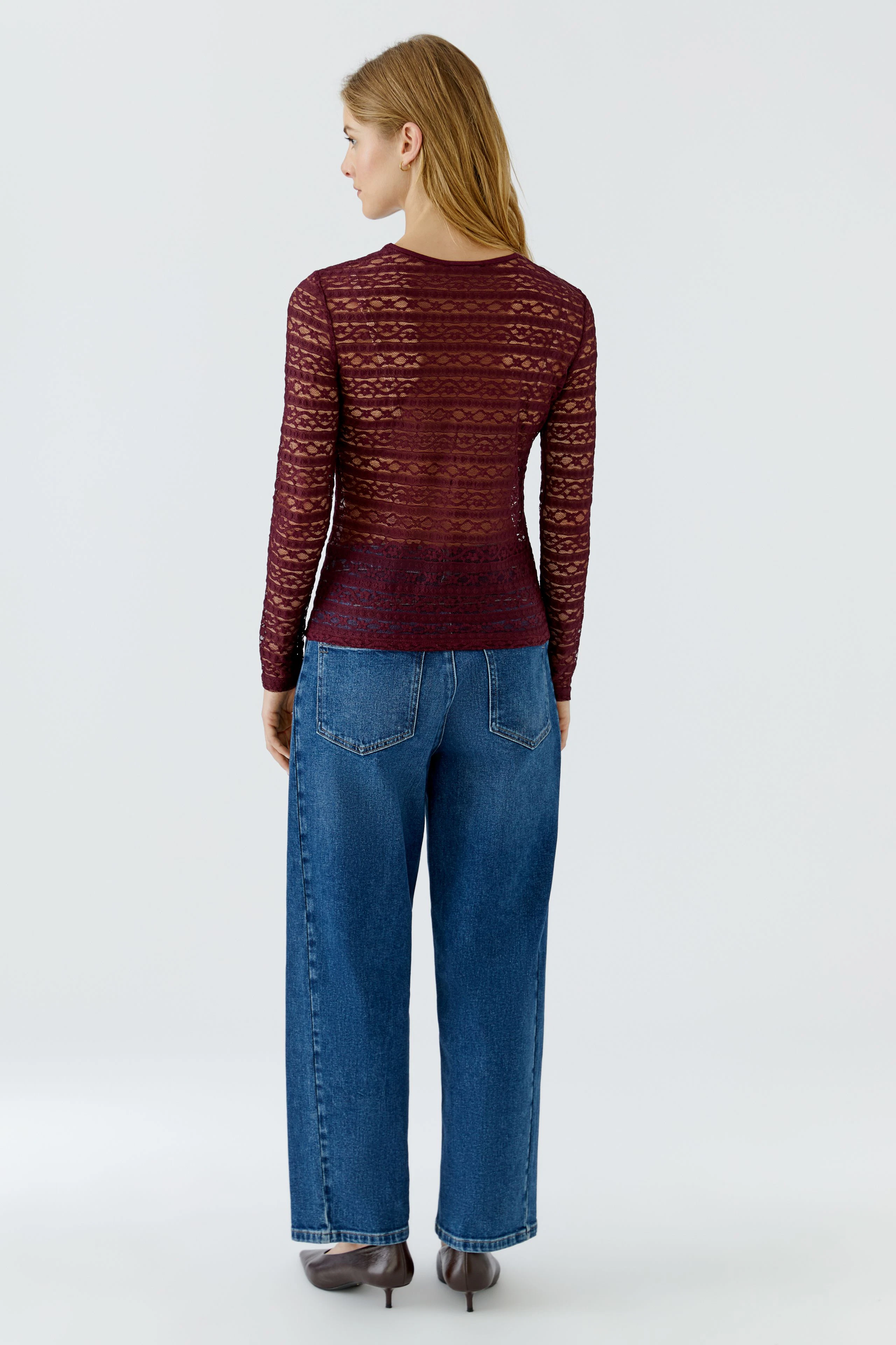 Frau im roten Langarmshirt aus Spitze, kombiniert mit blauen Jeans, von hinten gesehen.