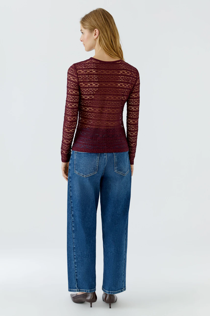 Frau im roten Langarmshirt aus Spitze, kombiniert mit blauen Jeans, von hinten gesehen.