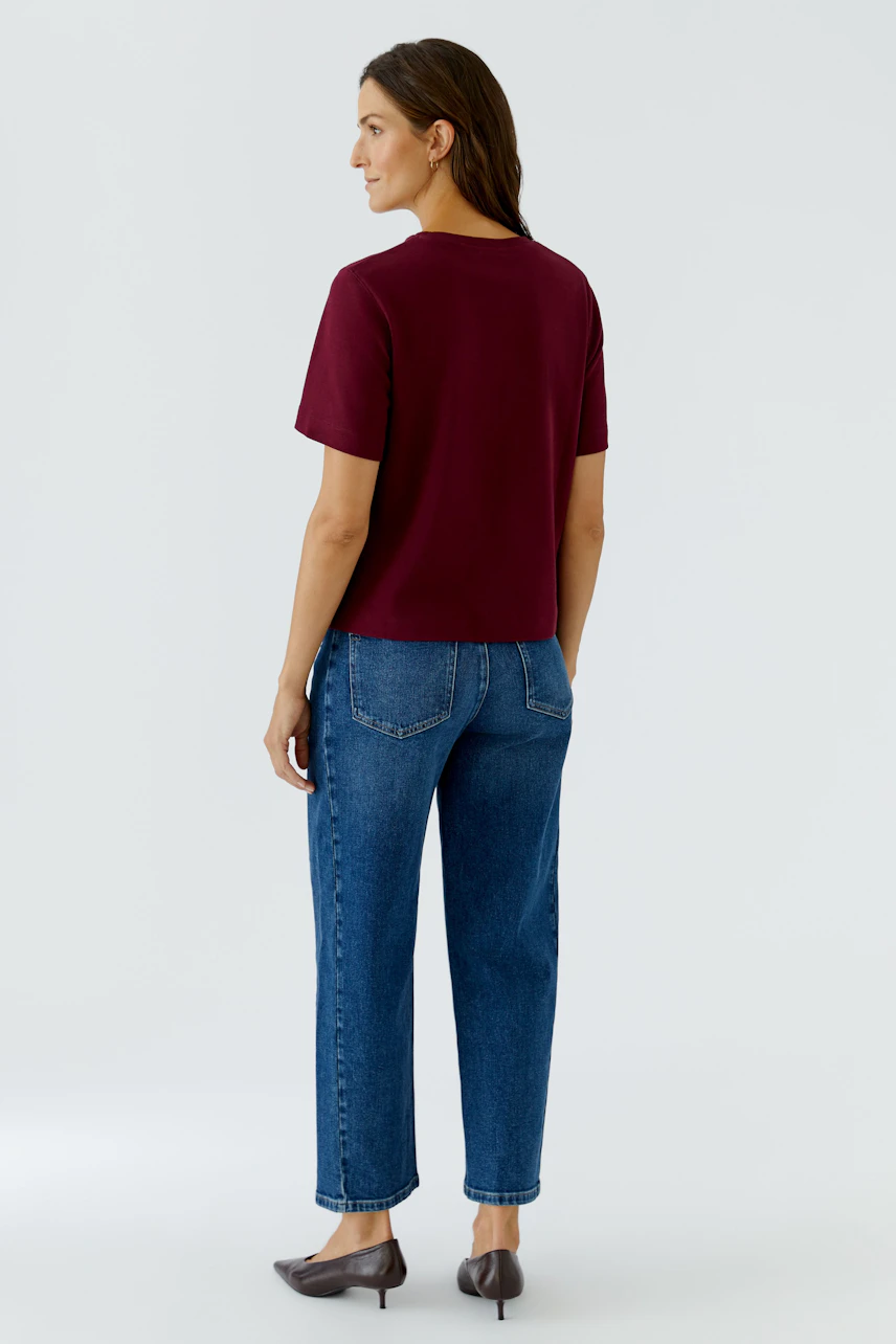 Frau im weinroten T-Shirt und blauer Jeans, von hinten gesehen. Lässiger Look mit flachen Schuhen.