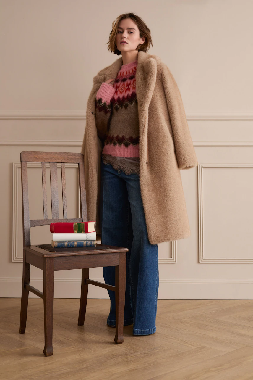 Winter coat: Woman in a cozy beige coat over a colorful pullover and jeans, standing confidently.