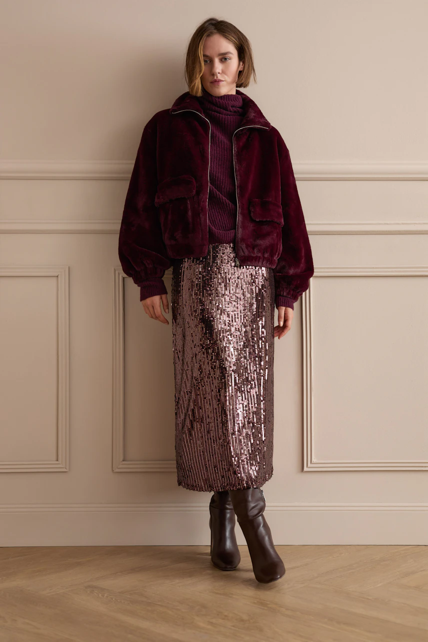 Woman in a sequin pencil skirt, burgundy jacket, and boots, standing confidently against a neutral wall.