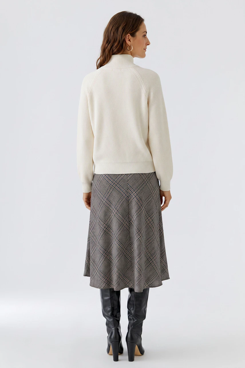 A-line skirt: Woman in a plaid A-line skirt, cream pullover, and knee-high boots, viewed from the back.